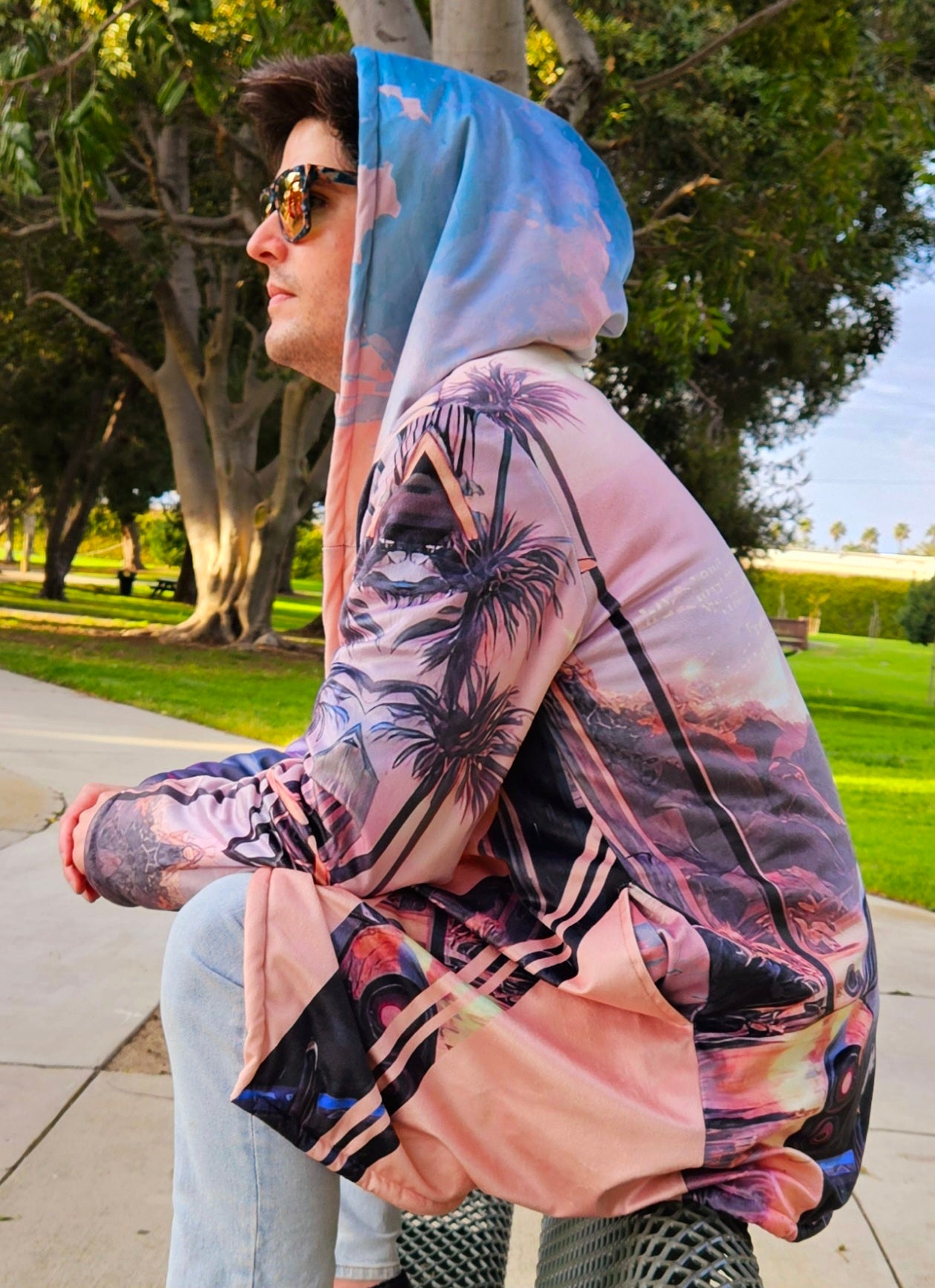 A man wearing sunglasses and a hoodie cloak sits outdoors, showcasing street fashion. His shirt features a palm tree design, emphasizing a casual, trendy look.